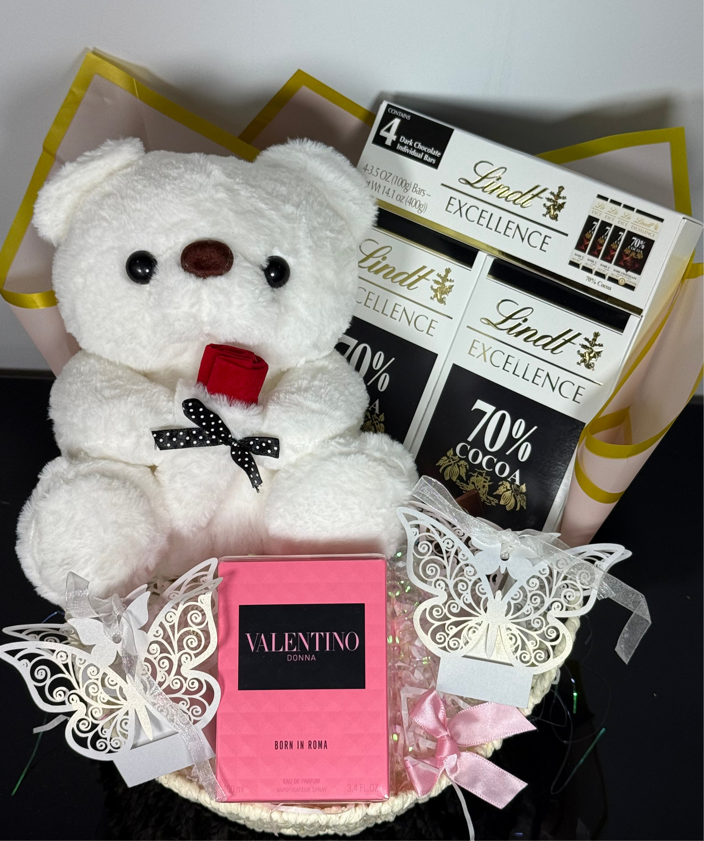 White teddy bear with a red bow, Lindt chocolate bars, and Valentino perfume in a decorative box.
