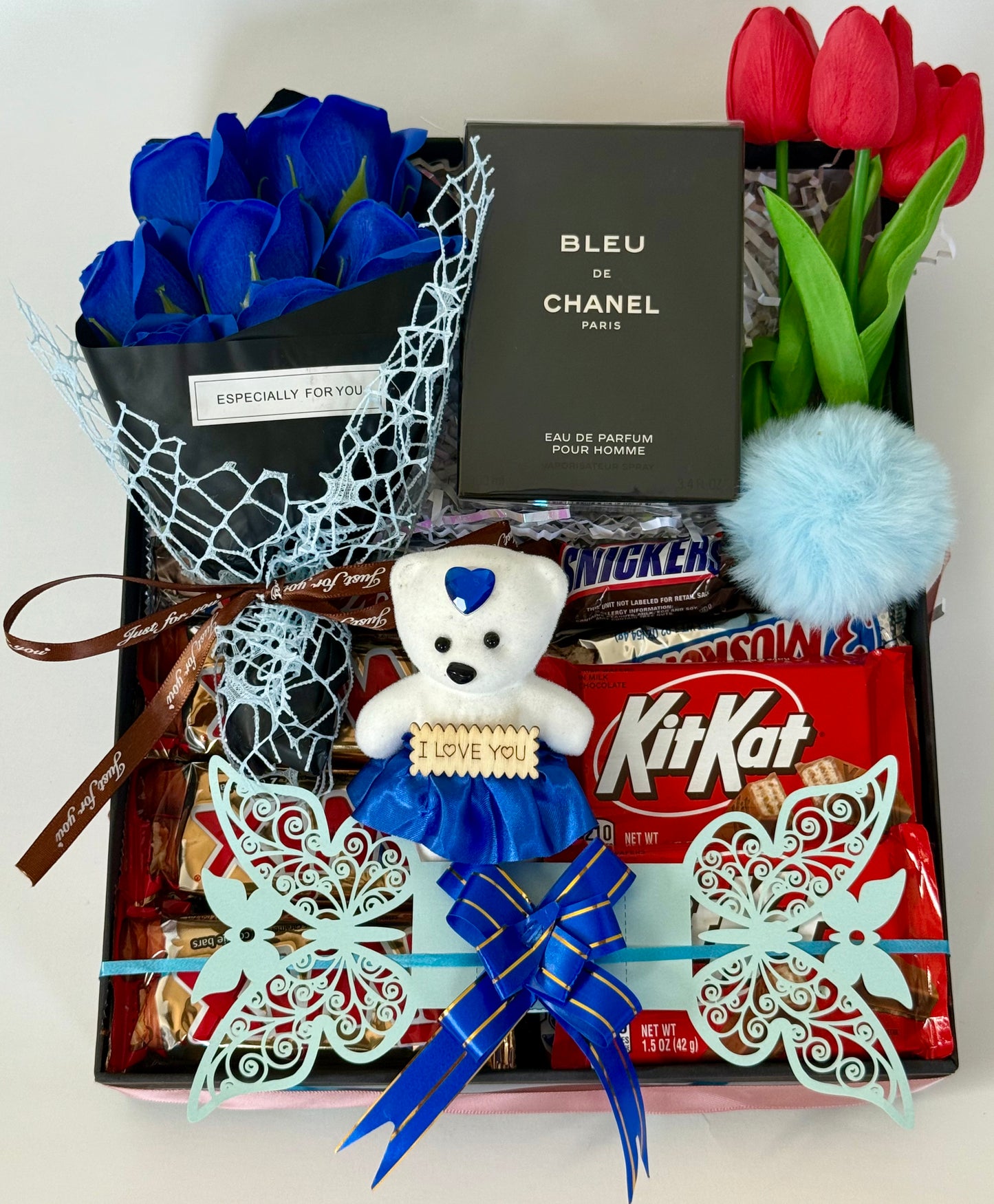 Gift basket with blue roses, teddy bear, KitKat bars, and Chanel perfume on a white background