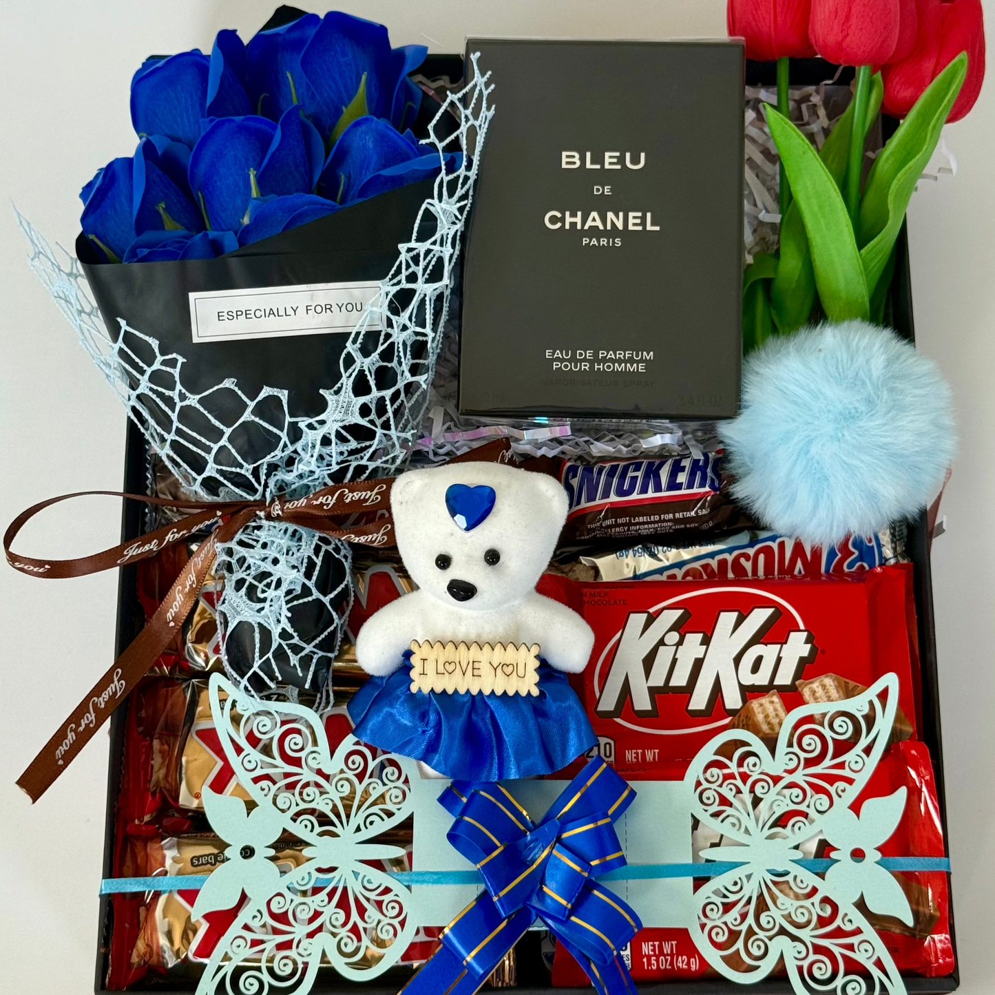 Gift basket with blue roses, teddy bear, KitKat bars, and Chanel perfume on a white background