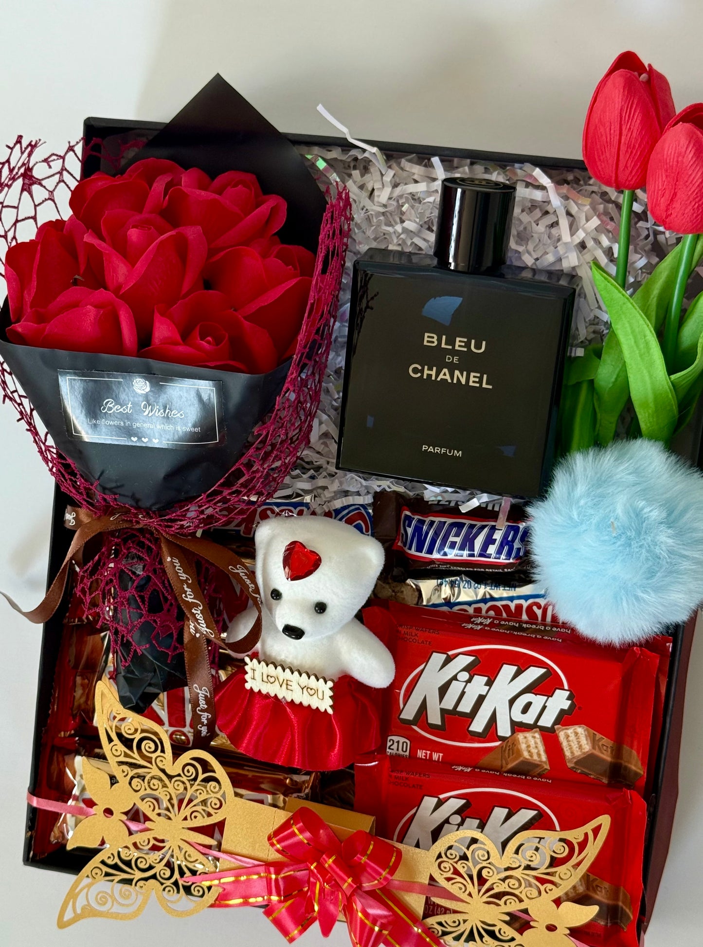 Gift box with red roses, Chanel perfume, teddy bear, KitKat chocolate bars, and tulips.