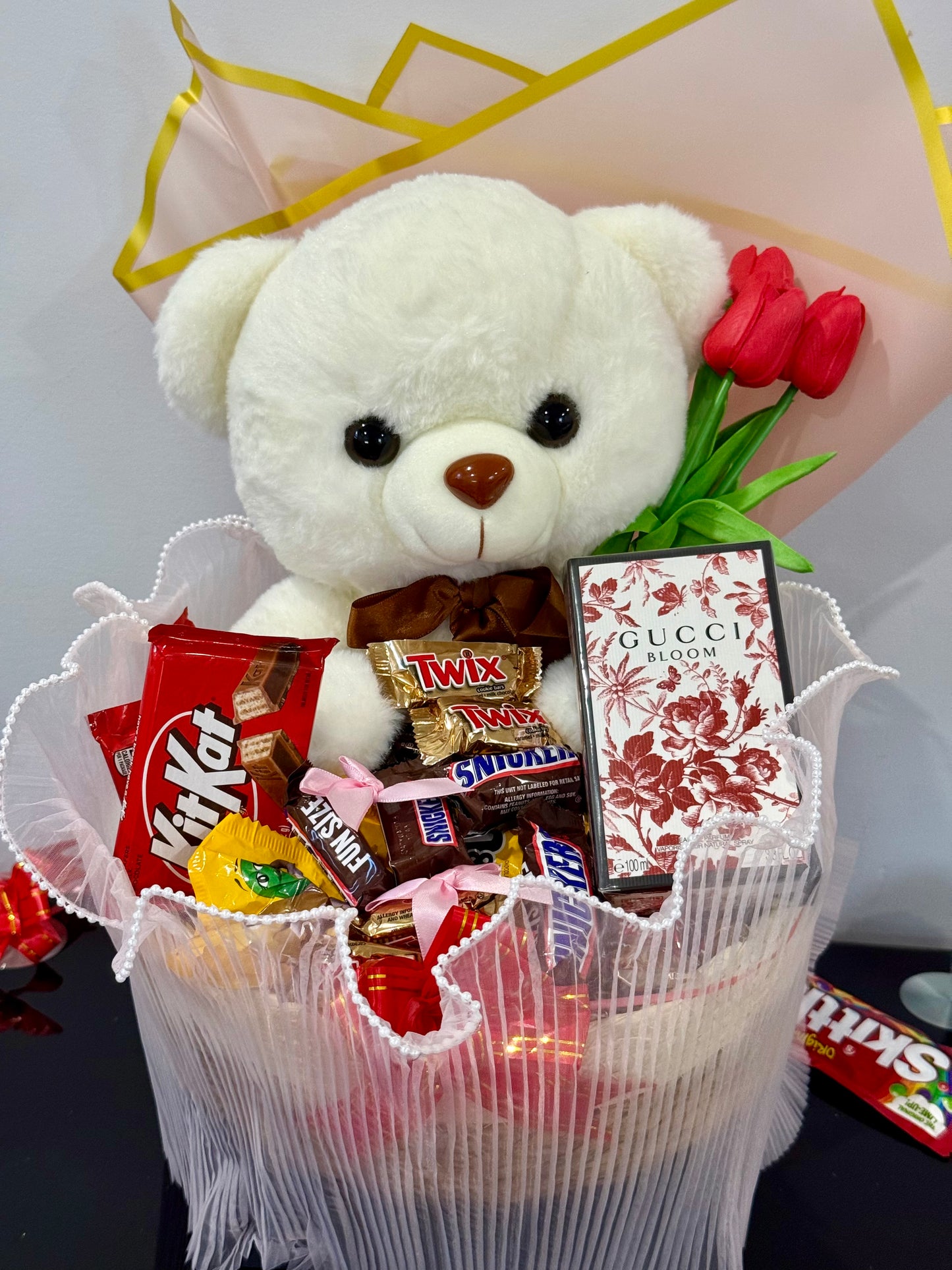White teddy bear with a bouquet of flowers and candy bars in a decorative basket.