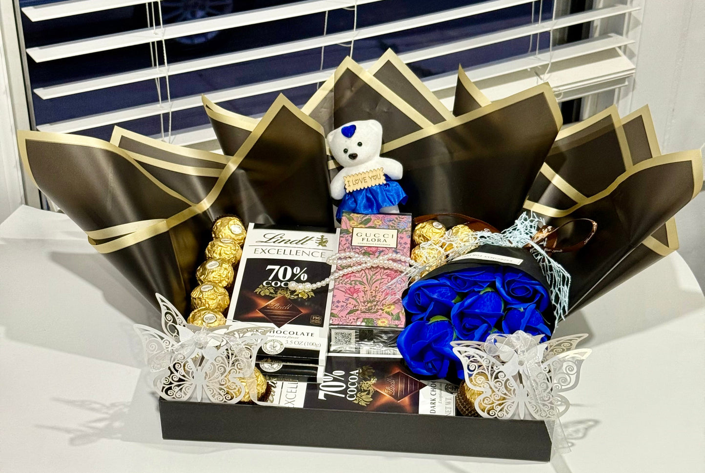 Gift basket with chocolates, flowers, and a teddy bear on a table with blinds in the background.
