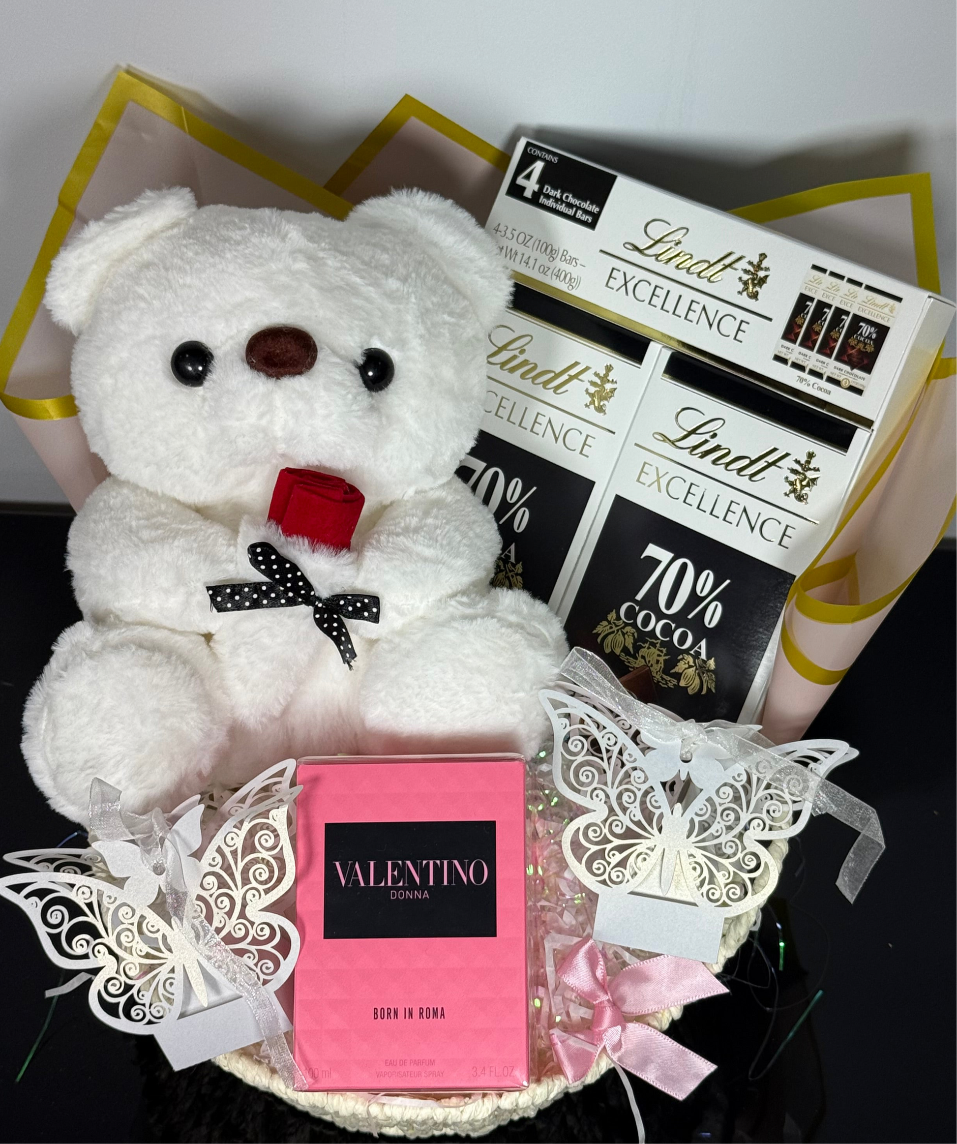 White teddy bear with a red bow, Lindt chocolate bars, and Valentino perfume in a decorative box.
